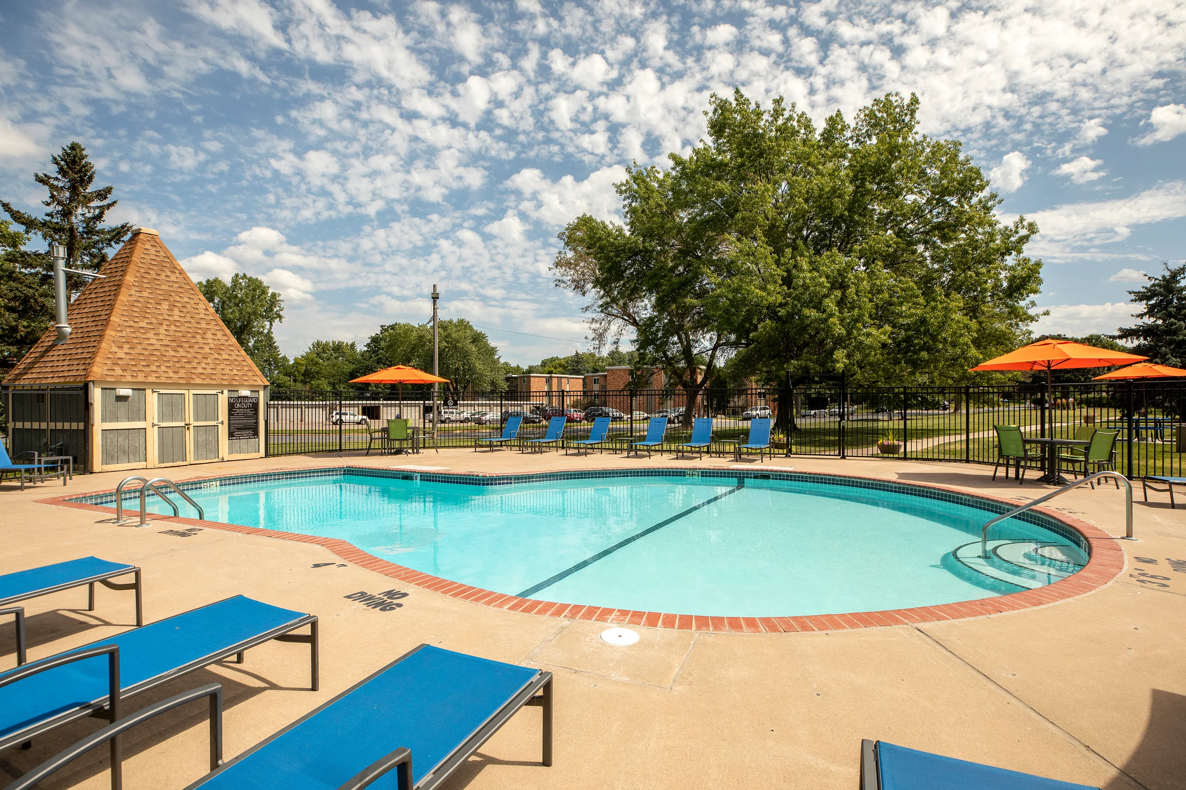 A sunny outdoor pool area featuring a circular swimming pool, surrounded by blue lounge chairs and orange umbrellas. Lush green trees are in the background under a blue sky with scattered clouds.
