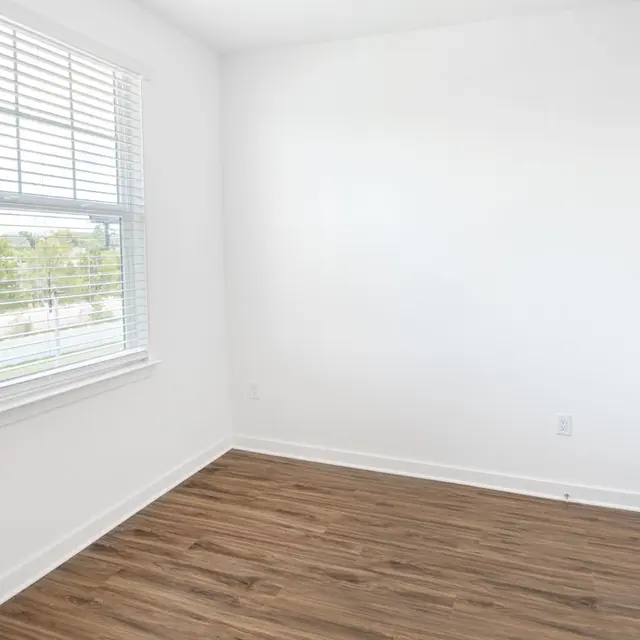 A bright, empty room with white walls and wooden flooring. It features a large window with blinds letting in natural light.