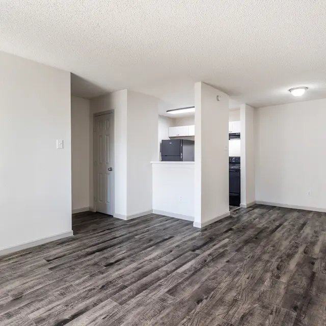 A spacious and empty apartment living area with wooden flooring and light-colored walls. There is a kitchen area visible in the background, featuring a dark colored appliance setup.
