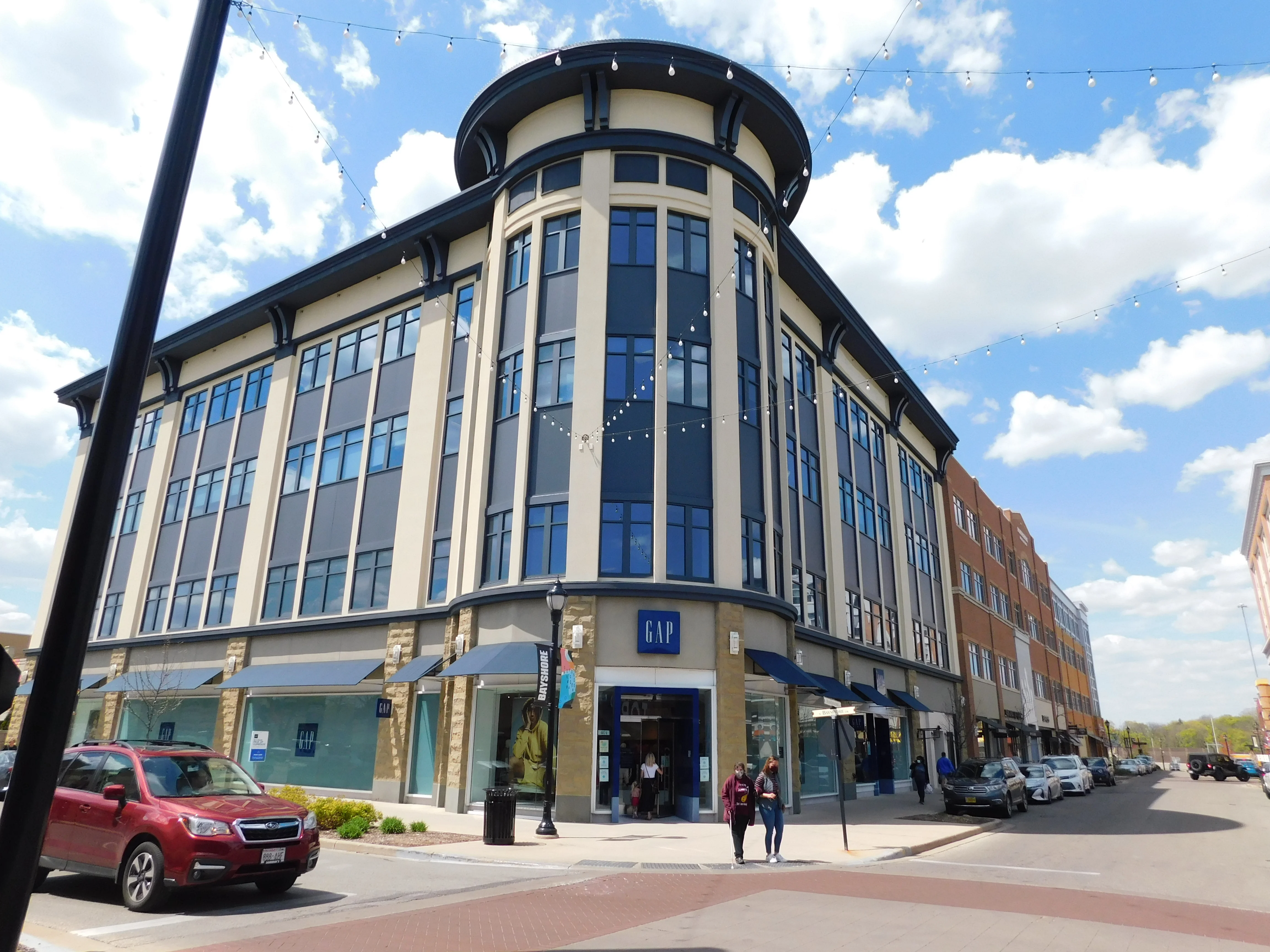 A modern multi-story building with a Gap store on the ground floor, surrounded by a busy street and other commercial buildings.