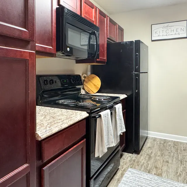 A modern kitchen featuring dark wood cabinets, a black stove, a black microwave, a black refrigerator, and a granite countertop. A cutting board and towels are visible near the stove, and a kitchen rug is on the floor.