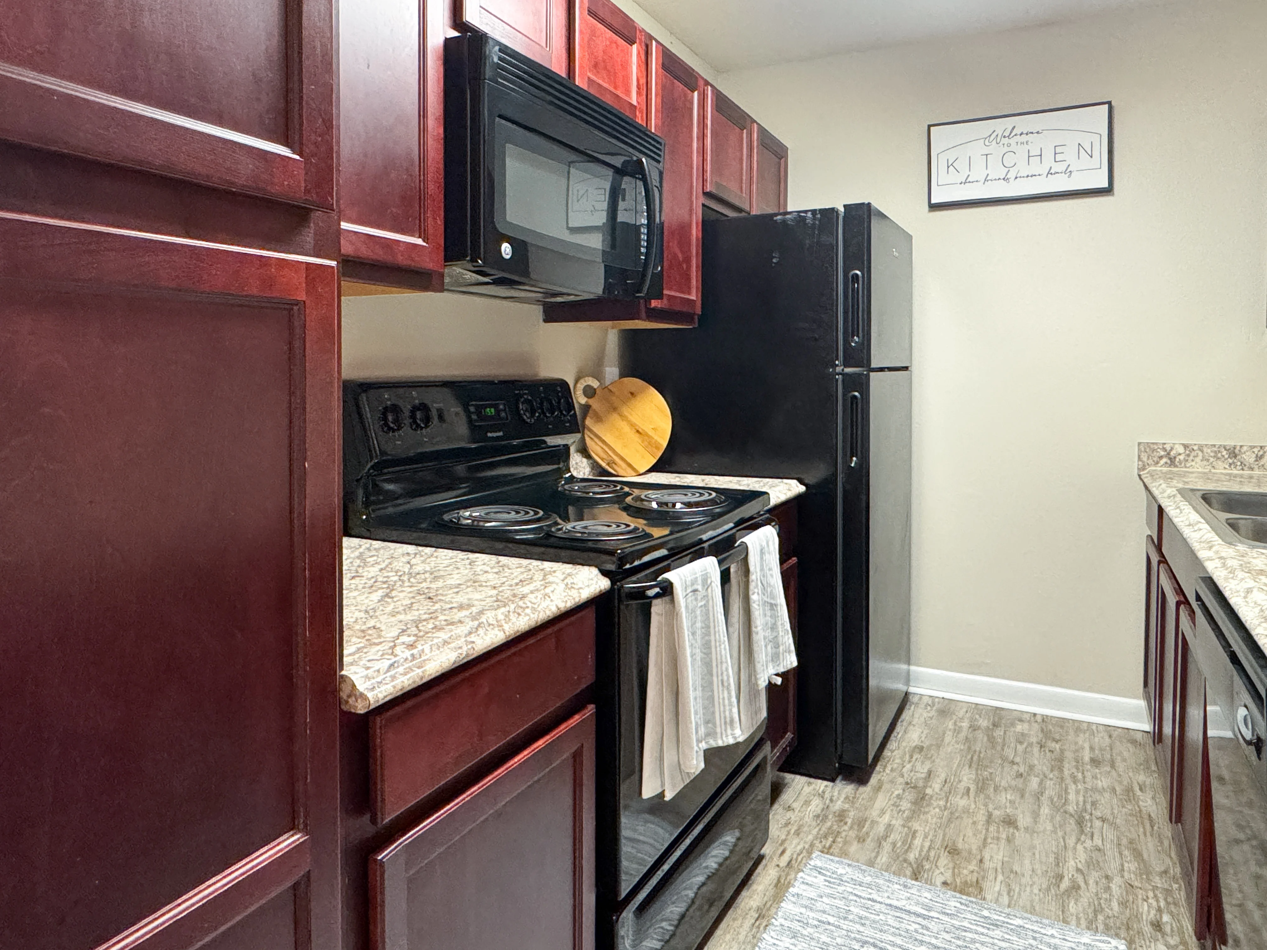 A modern kitchen featuring dark wood cabinets, a black stove, a black microwave, a black refrigerator, and a granite countertop. A cutting board and towels are visible near the stove, and a kitchen rug is on the floor.