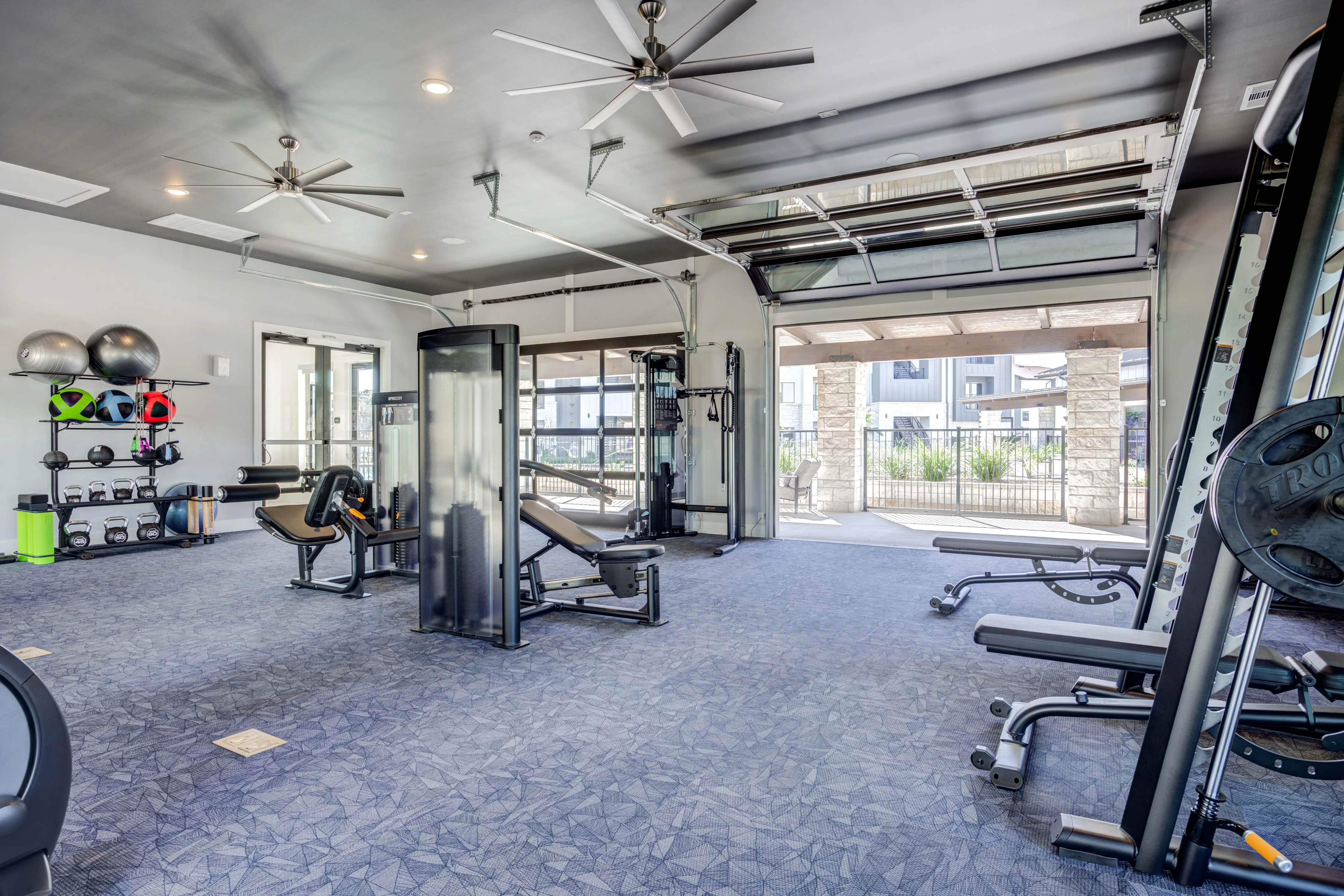 Interior view of a modern gym with various fitness equipment, large windows, and a view of the outdoors.