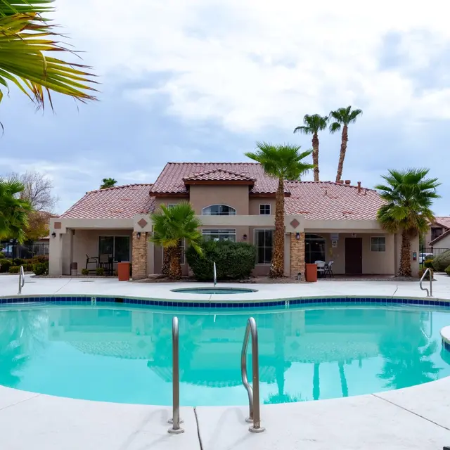 A serene pool area featuring a large, oval swimming pool surrounded by palm trees and well-maintained landscaping. In the background, a spacious building with a red tiled roof is visible under a cloudy sky.