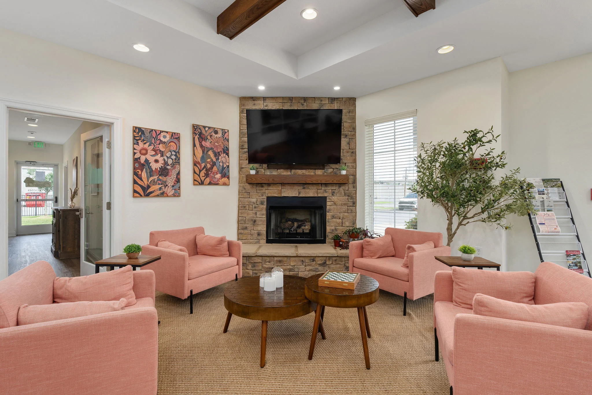 A cozy community lounge area featuring pink armchairs, a stone fireplace, a mounted TV, and a decorative plant.