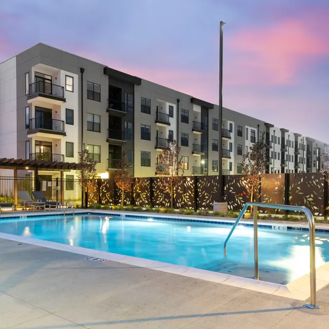 Modern apartment complex with a swimming pool at dusk, featuring lounge chairs and decorative fencing.
