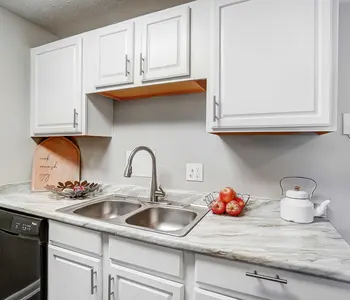 A modern kitchen with white cabinets, a dark dishwasher, a marble countertop, a sink with a faucet, a bowl of apples, and a tea kettle.