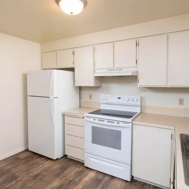 An interior view of a clean, modern kitchen featuring a white refrigerator, white stove, and light-colored cabinets. The flooring is a wood-like laminate, and there is a double sink on the right.