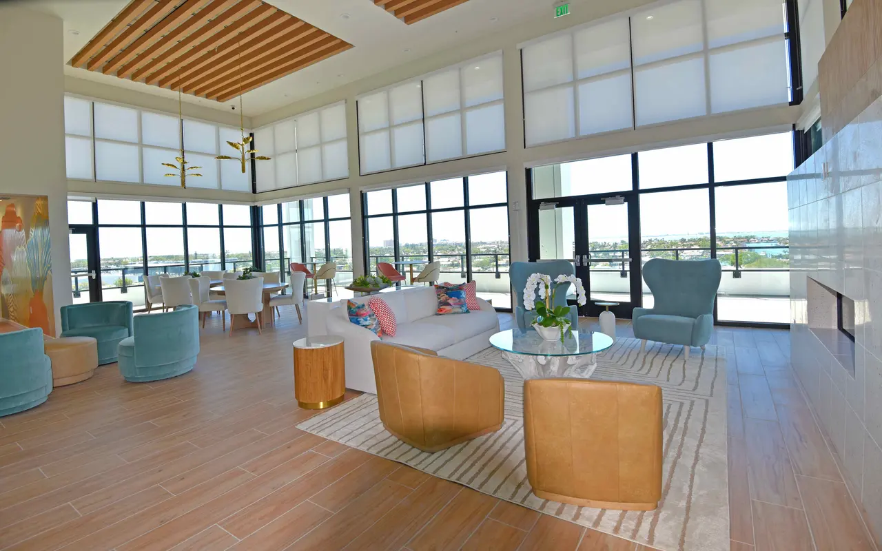 A spacious modern living area with large windows providing ample natural light, featuring stylish furniture including a white sofa, colorful armchairs, and a round glass table, with decorative plants and a cozy rug.