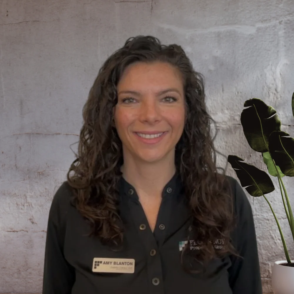 A woman with curly hair smiles in front of a textured wall and a plant.