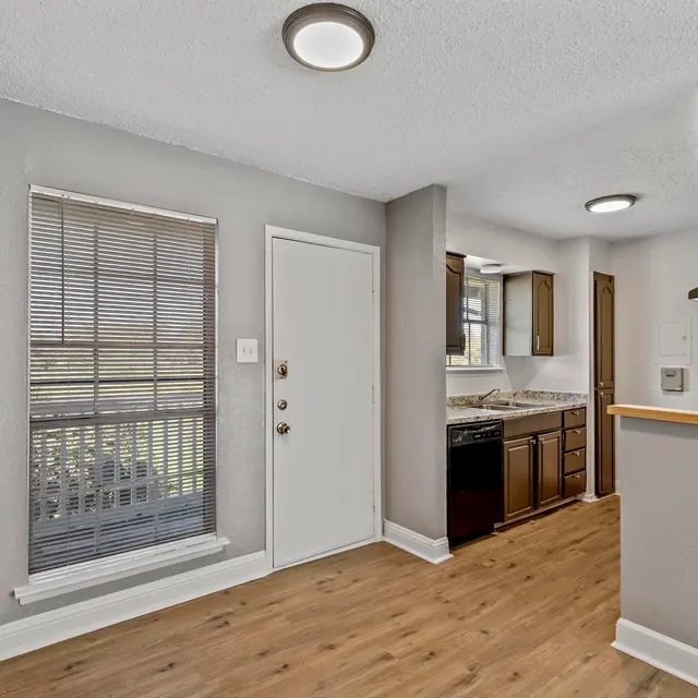 View of a modern apartment interior with a kitchen and living area. The space features wood flooring, a window with blinds, and dark wooden cabinets.