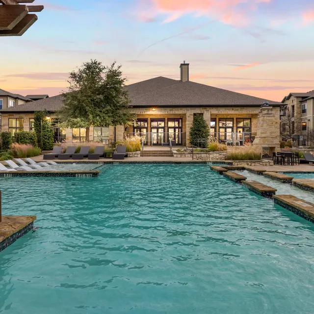 A swimming pool area featuring a sparkling blue pool, surrounded by lounge chairs and tables, with a clubhouse in the background. The sky shows soft pastel colors of sunset.