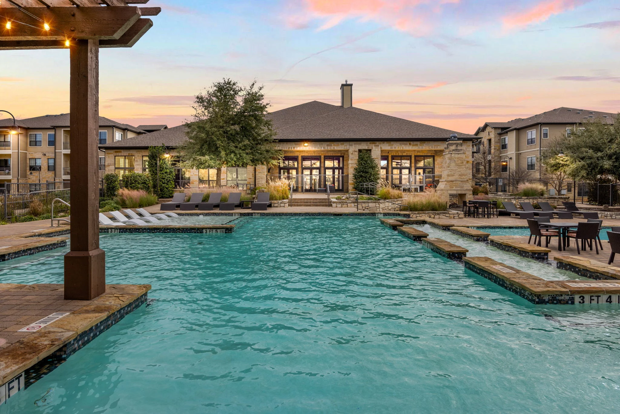 A swimming pool area featuring a sparkling blue pool, surrounded by lounge chairs and tables, with a clubhouse in the background. The sky shows soft pastel colors of sunset.