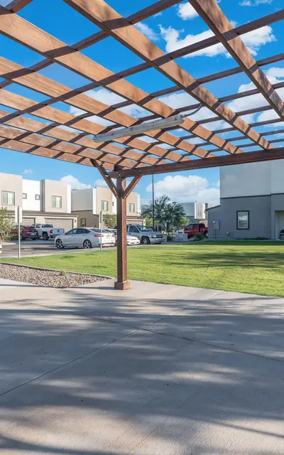 Outdoor Pergola and Lawn Area An outdoor pergola with a wooden structure, surrounded by grass and modern buildings in the background, under a clear blue sky with few clouds.