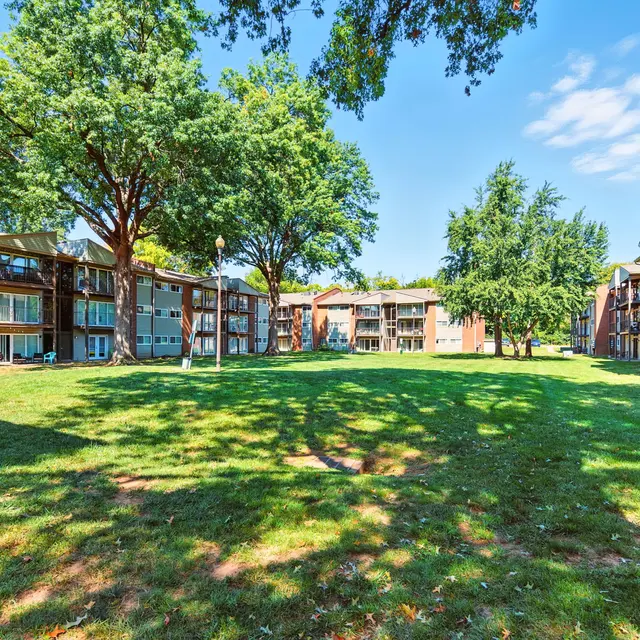 A well-maintained apartment complex surrounded by trees and a spacious green lawn under a clear blue sky.