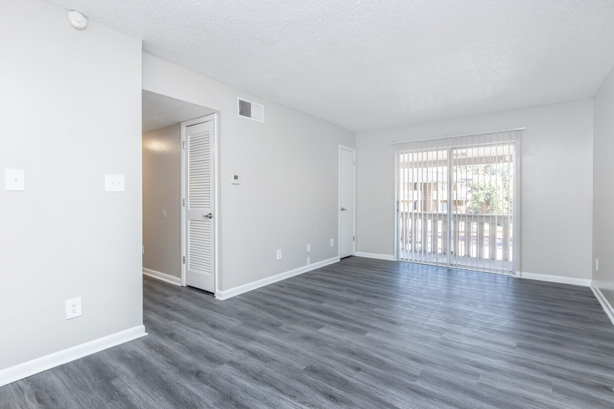 Modern Empty Living Room A spacious, empty living room featuring light gray walls and modern laminate flooring. There is a sliding glass door leading to a small balcony, and a couple of doors on the left side, one of which likely leads to a closet. The room is well-lit, emphasizing its open layout.