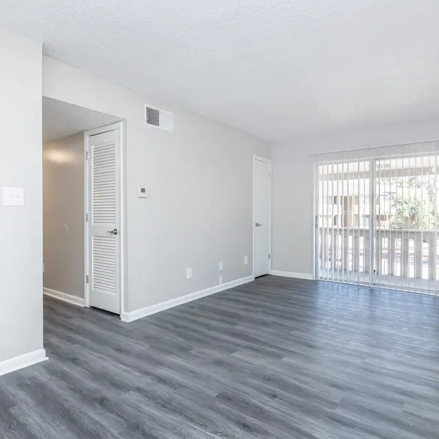 A spacious, empty living room featuring light gray walls and modern laminate flooring. There is a sliding glass door leading to a small balcony, and a couple of doors on the left side, one of which likely leads to a closet. The room is well-lit, emphasizing its open layout.