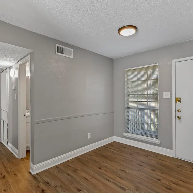 A well-lit living room area with gray walls, a window, and a door leading outside. The floor features light brown laminate, and there is a small hallway visible leading to another room. The ceiling has a circular light fixture.