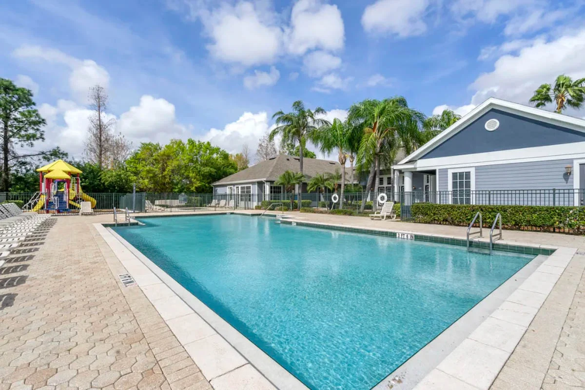 A clear swimming pool surrounded by landscaped greenery and a play structure nearby under a blue sky.
