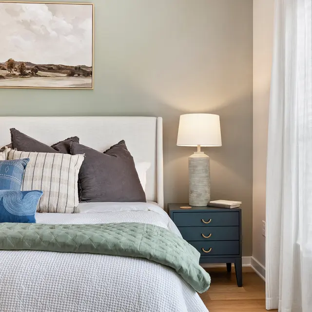 A modern bedroom featuring a white bed with various patterned pillows, a bedside lamp on a blue nightstand, and a wall-mounted painting. Bright sunlight filters through a window with white curtains.
