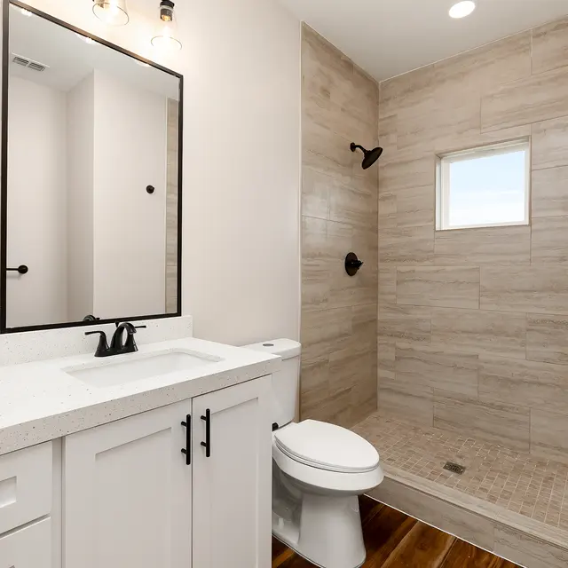 A modern bathroom featuring a white vanity with a sink and faucet, a large mirror above the vanity, a toilet, and a walk-in shower with tiled walls and a small window for natural light.