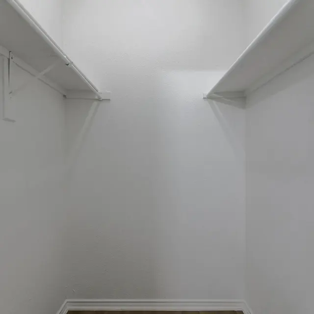 An empty walk-in closet with white walls and wooden floor, featuring two parallel shelves.