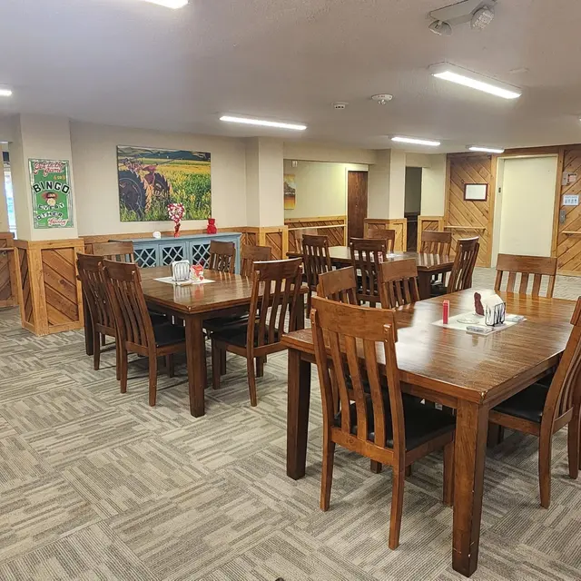 A spacious dining area with several wooden tables and chairs, carpeted flooring, and a few blue upholstered chairs. The walls have wooden paneling and a colorful painting is displayed on one wall.