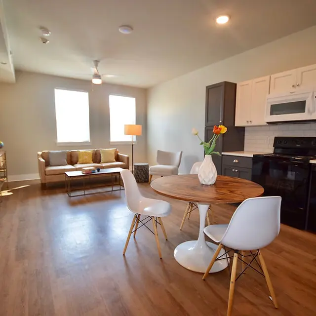 A modern apartment interior featuring a living area and kitchen with white cabinets and dark appliances, a round dining table with white chairs, and natural light from large windows.