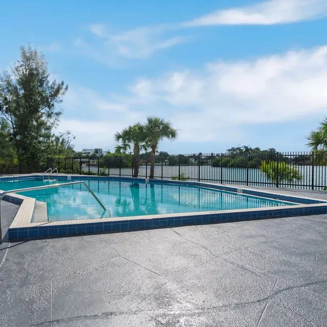 A serene swimming pool surrounded by a black fence, with a view of a lake and landscaped greenery in the background under a blue sky.