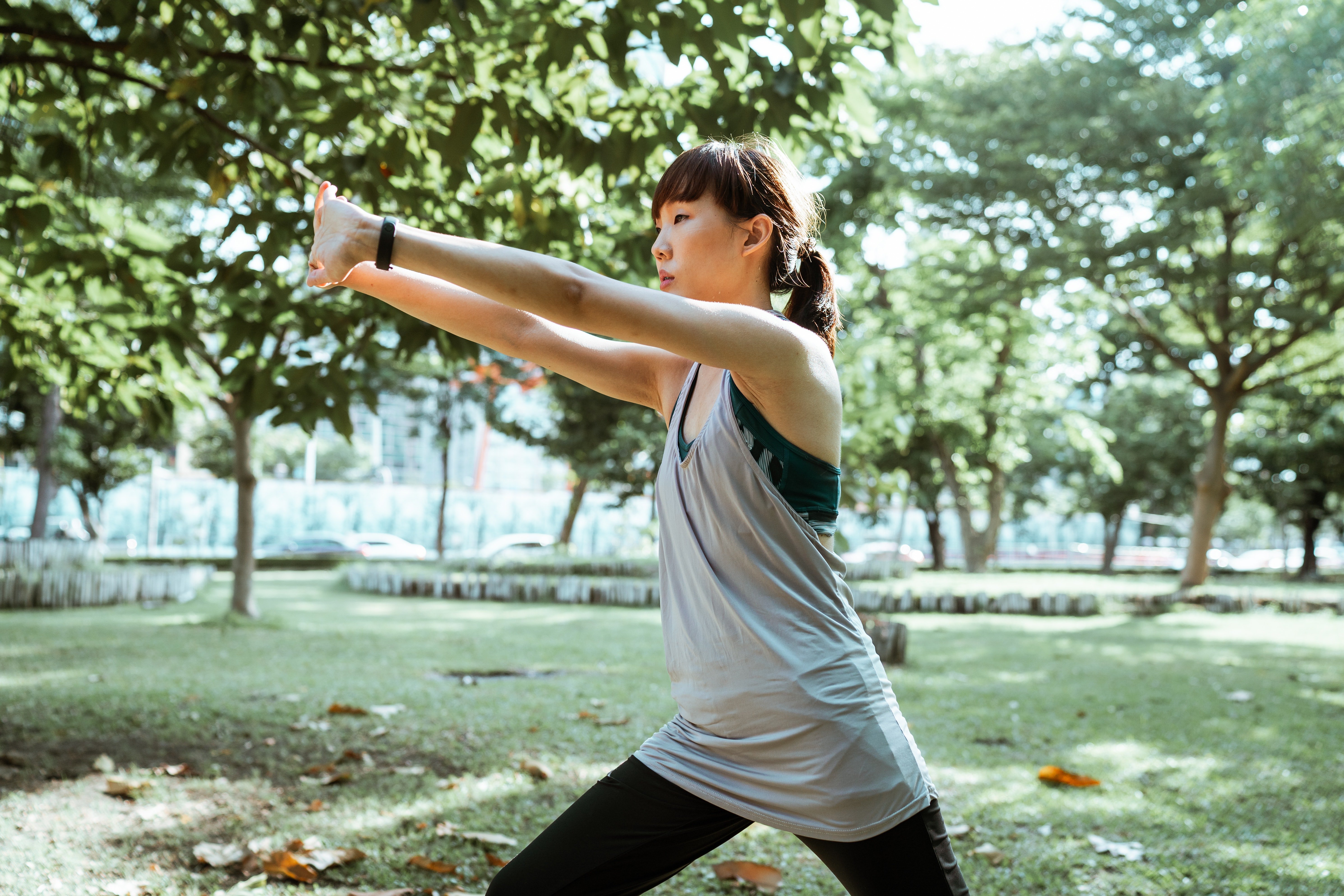 A woman performing an exercise in a park, stretching her arms forward while standing on one leg in a lunge position. The background features lush green trees and a grassy area.
