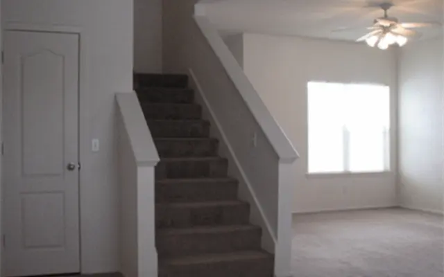 Living Room with Staircase Interior view of a living room featuring a staircase leading to the upper floor. A large window allows natural light to enter the room, which has a carpeted floor and neutral-colored walls.