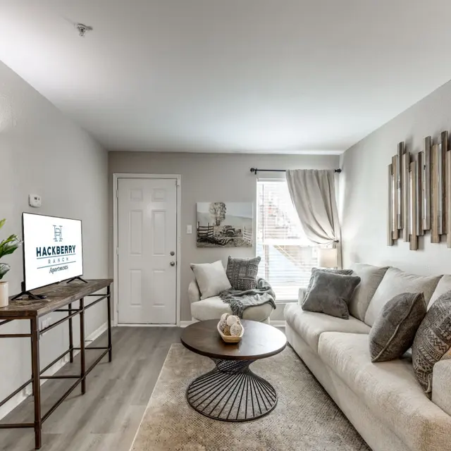 A contemporary living room featuring a light-colored sofa, decorative pillows, a round coffee table, and a television on a stand. Natural light enters through a window adorned with curtains, and a wall decor piece made of wood is visible.