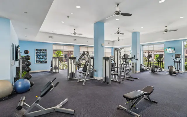 A spacious gym interior featuring various exercise equipment including weight machines, benches, and cardio machines. The room is brightly lit with large windows allowing natural light in, and has a blue color scheme with white accents.