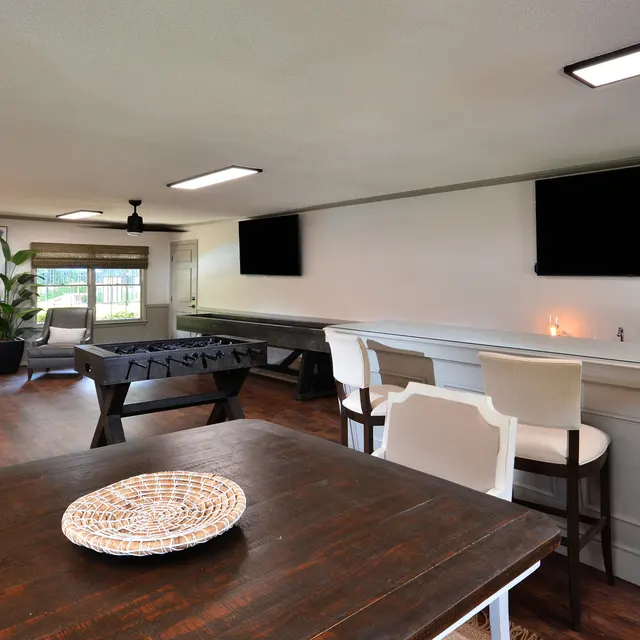 A modern living space featuring a dining table, lounge area with comfortable chairs, a foosball table, and two flat-screen TVs mounted on the wall.