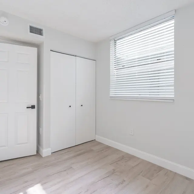 A well-lit empty room with light grey walls and wooden floors, featuring a closed white double-door closet and a window with blinds.