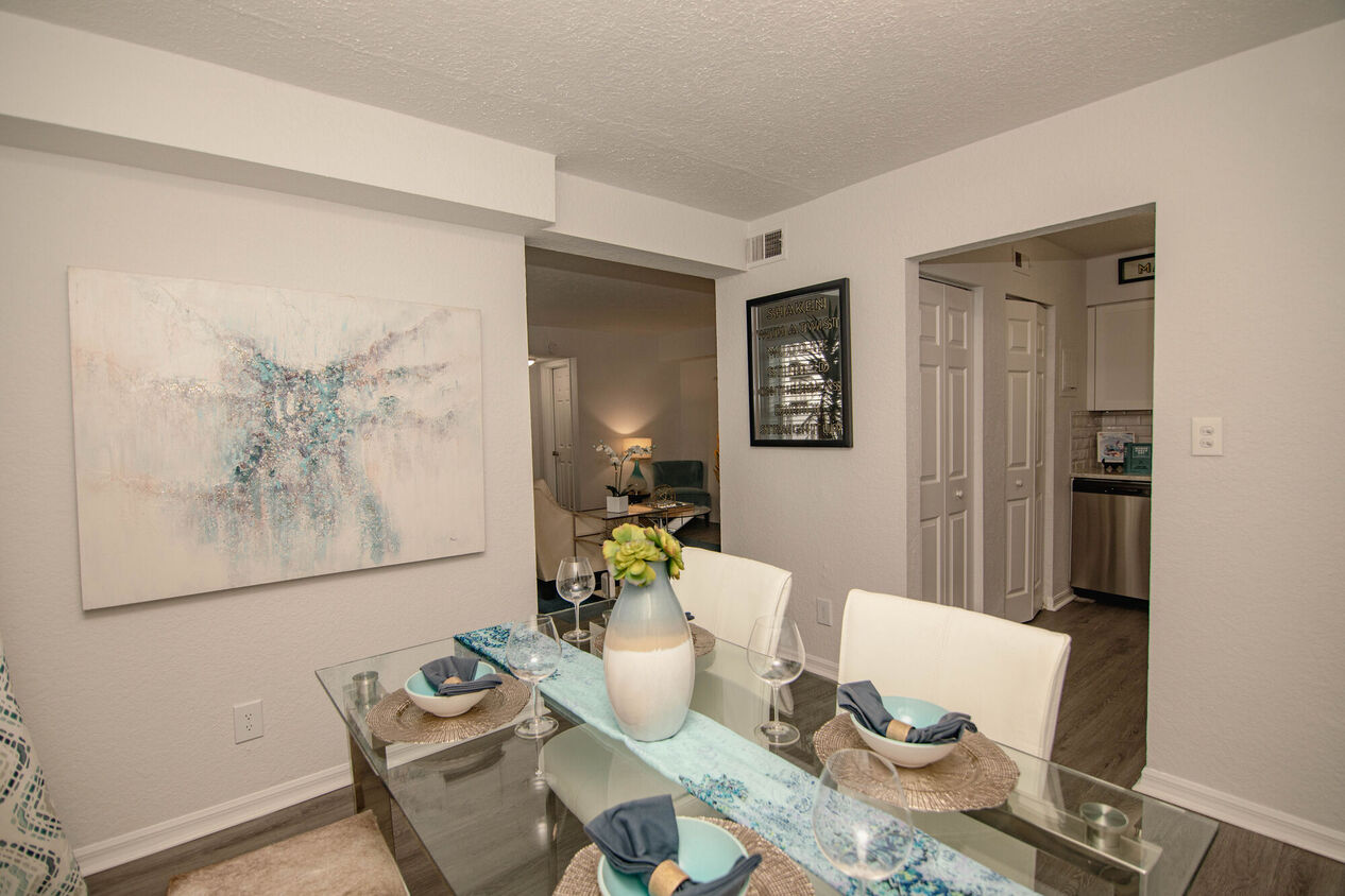 A modern dining area with a glass table set for a meal, featuring white chairs and a decorative vase with flowers. A colorful artwork is positioned on the wall, and the layout opens to a living space beyond.