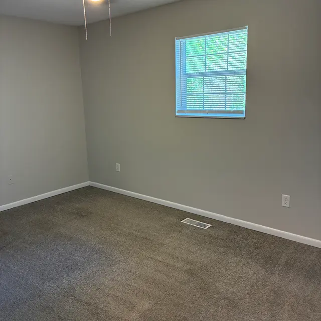 Empty Room with Ceiling Fan and Window A spacious empty room with light gray walls and carpet. The room features a ceiling fan and a window with blinds letting in natural light.