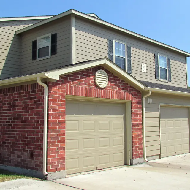 Exterior view of a two-story apartment building with a brick lower section and beige upper section, featuring garage doors and several windows on a sunny day.