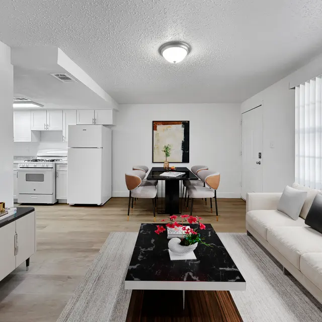 A modern living room featuring a white sofa, a black coffee table with flowers, a television, and a dining area with chairs in a well-lit space.
