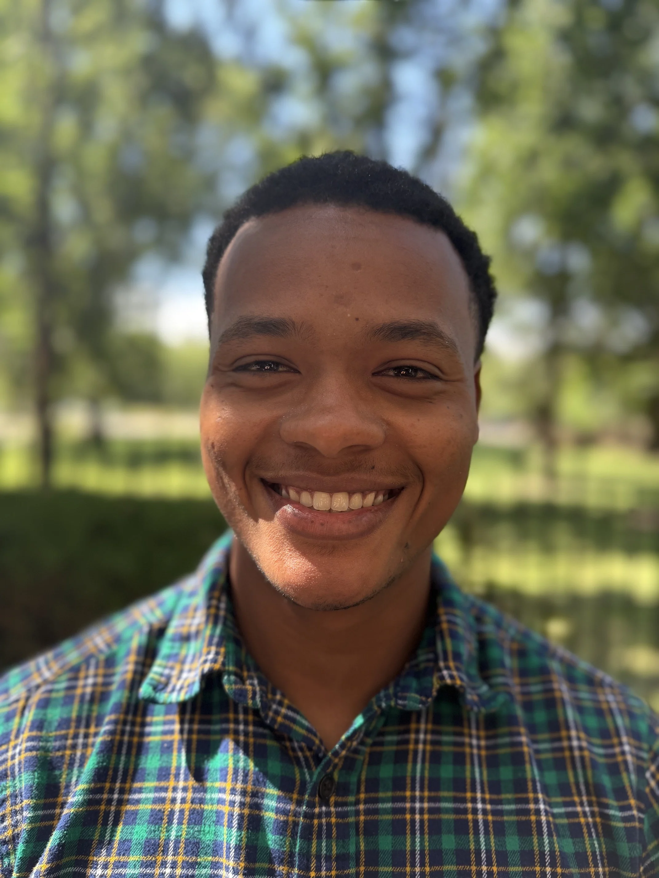 Smiling Young Man Outdoors A young man with a big smile standing in front of a green background, wearing a checkered shirt with shades of blue and green.