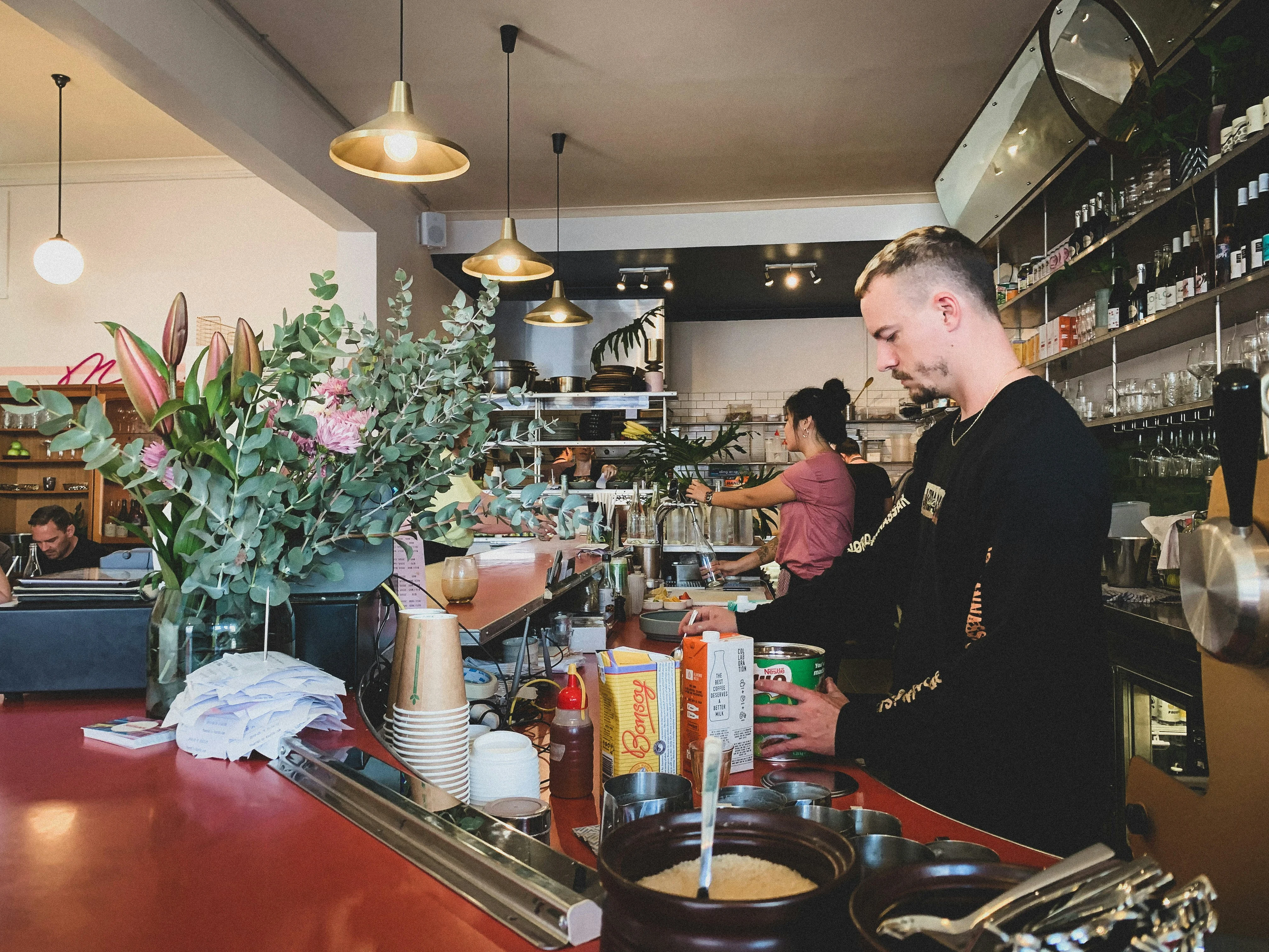 Café Interior with Barista A busy café interior with a male barista at the counter preparing drinks. Flowers adorn the counter, and shelves filled with drinks are visible in the background. Another person is engaged in a task in the back area.