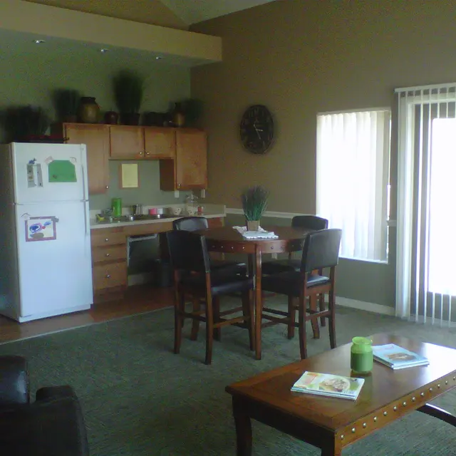 Cozy Living Space Interior An interior view of a cozy living space featuring a kitchenette, dining area, and seating area. The kitchenette has wooden cabinets and a fridge, while the dining area includes a table and chairs. Sunlight streams through sliding glass doors leading to the outside.