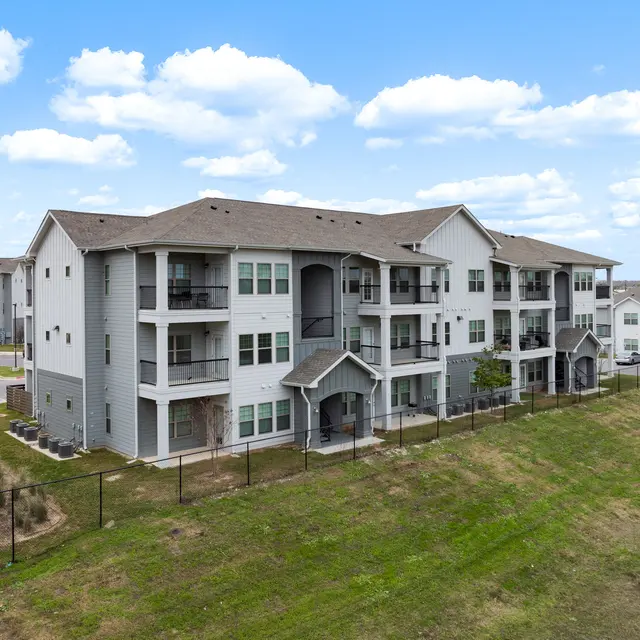 A modern apartment complex featuring multiple buildings with balconies and a grassy area nearby. The sky is clear with a few clouds.