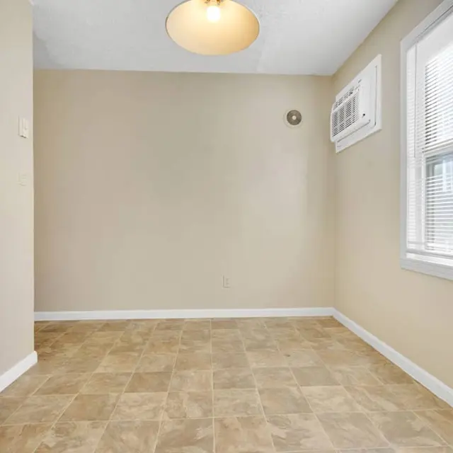 An empty room with a tiled floor, beige walls, and a window with blinds. There's an air conditioning unit under the window and a ceiling light fixture.