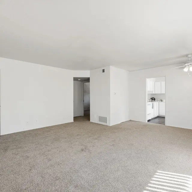 Spacious living room area in an apartment with beige carpet and white walls, featuring a door labeled 'HOME' and a visible kitchen area in the background.