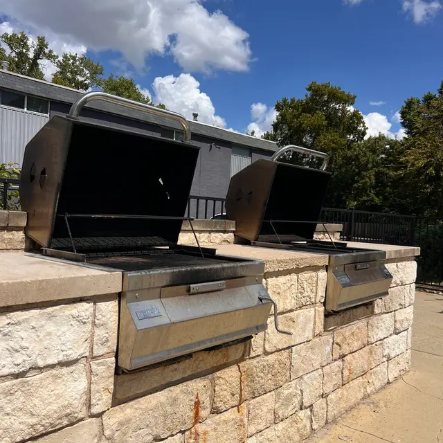 Outdoor Grills Two open outdoor grills beside a stone wall under a clear blue sky with fluffy white clouds.