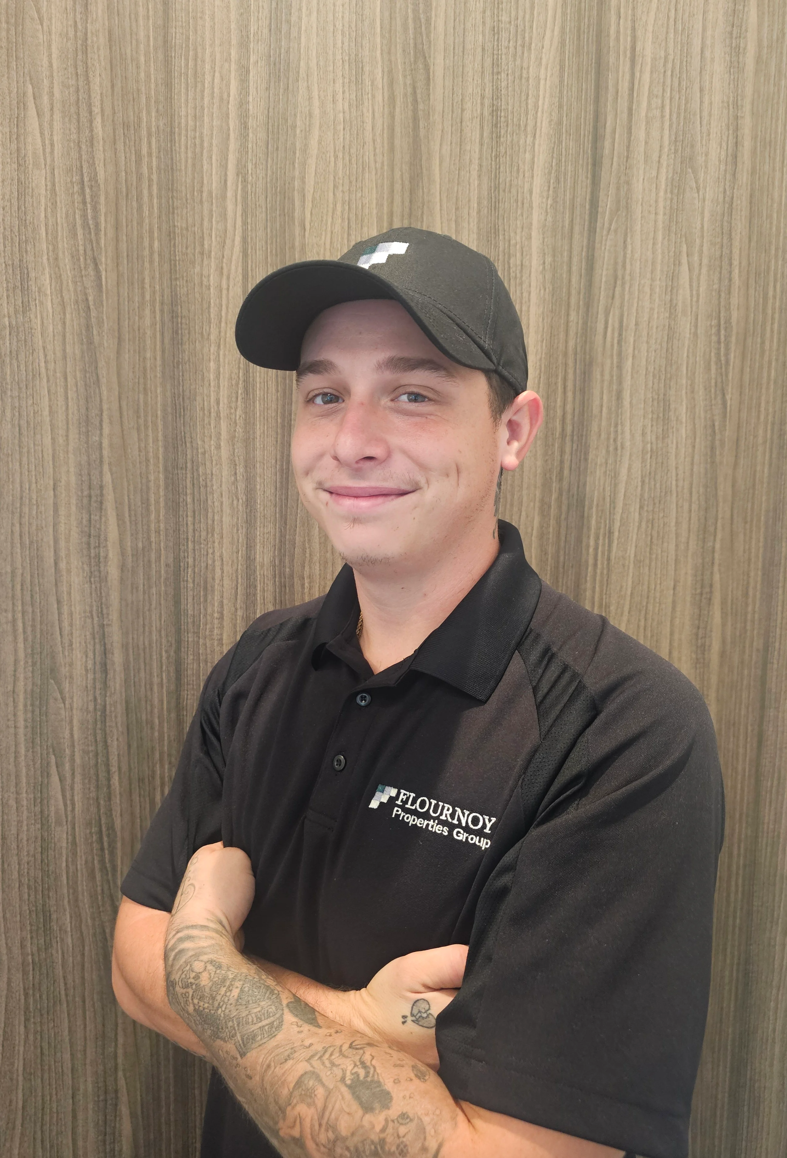 Employee of Flournooy Plumbing Group A young man with tattoos on his arm is standing in front of a wooden background. He is wearing a black cap and a black polo shirt with the logo of a company. He is smiling confidently with his arms crossed.