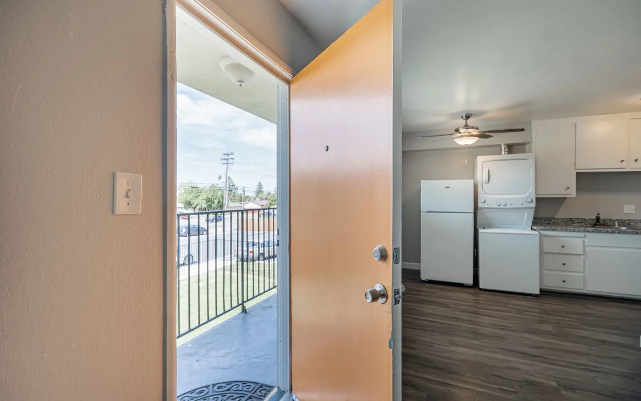 View of an apartment entryway with an open door, leading into a room featuring a kitchen and laundry area. A balcony and outdoor view with clear skies are visible through the doorway.