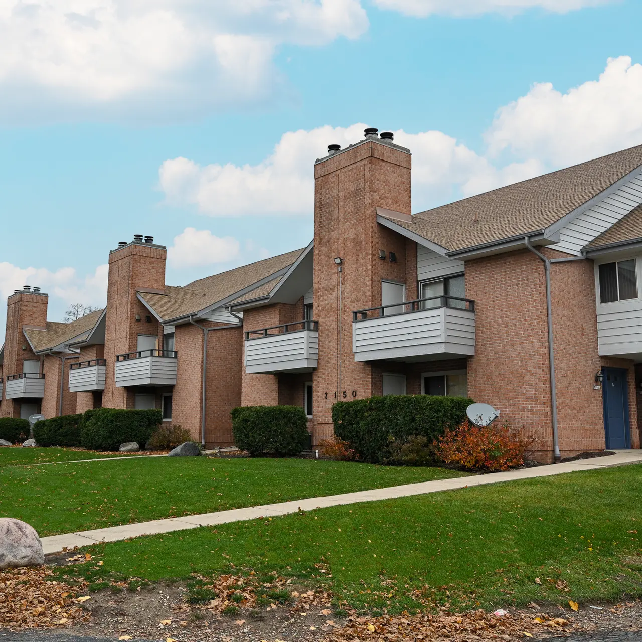 A brick apartment complex featuring two-story buildings with balconies, surrounded by a grassy area and a walkway. There are trees and shrubs in the vicinity, with a cloudy sky above.