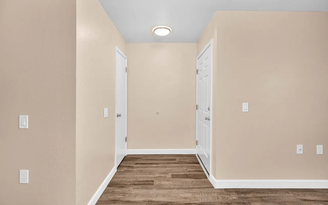 An empty room with light beige walls and wood flooring. There are two closed white doors on the left and right, and a ceiling light is visible above.